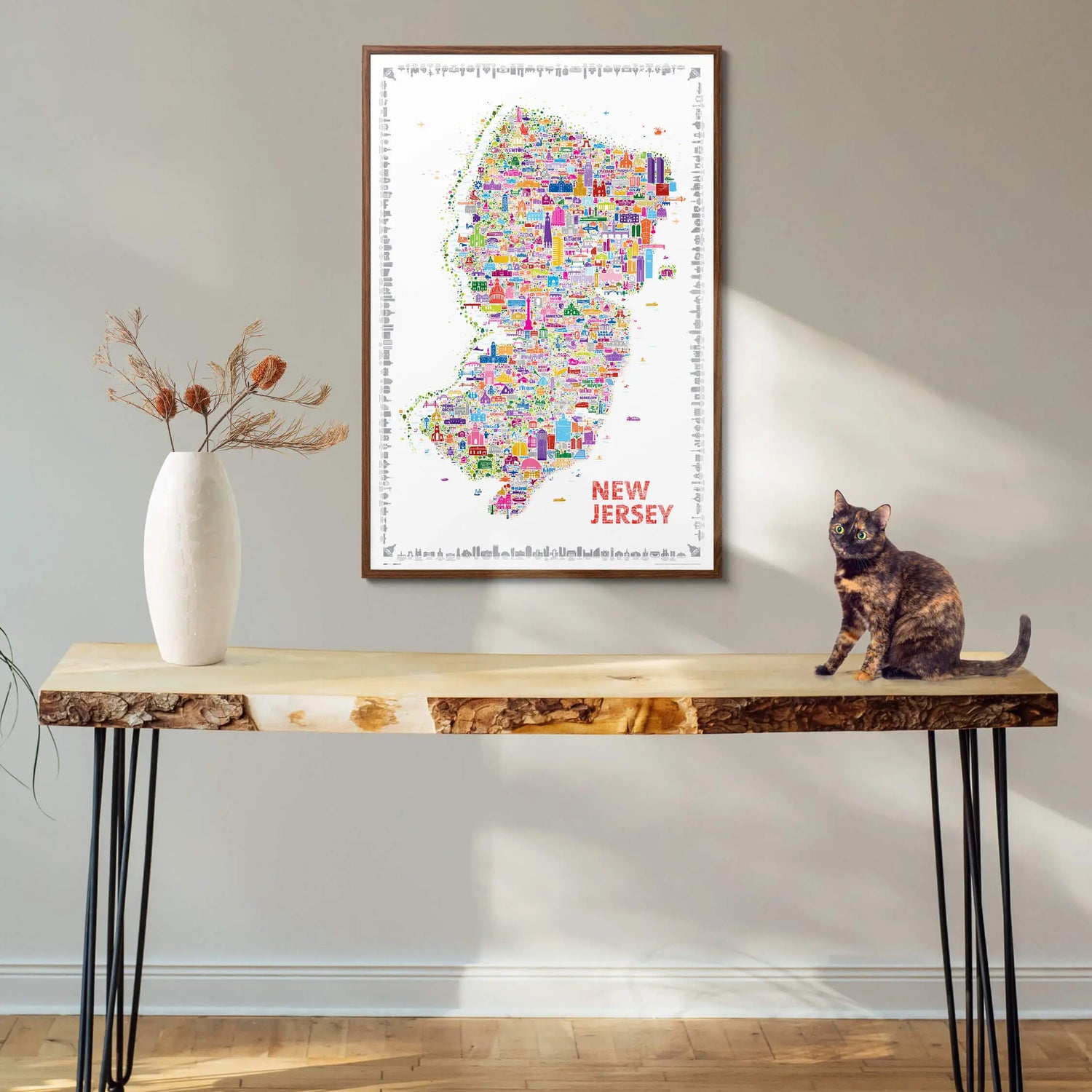Colorful New Jersey city map wall art framed in walnut wood, displayed above a rustic console table with a white vase and a cat sitting beside it — modern home decor by Alfalfa New York.