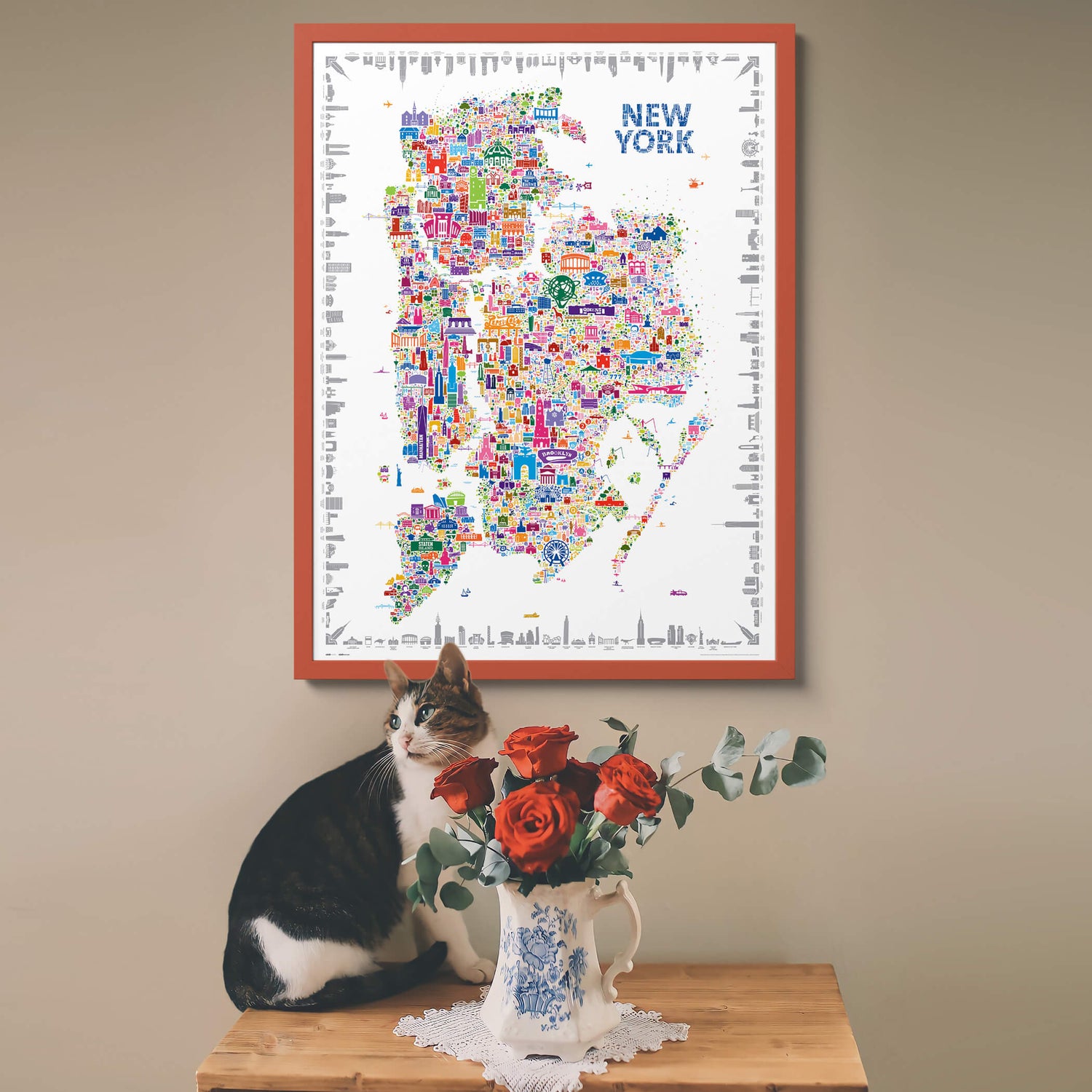 Colorful New York map poster on a wall with a cat and flowers on a table below.