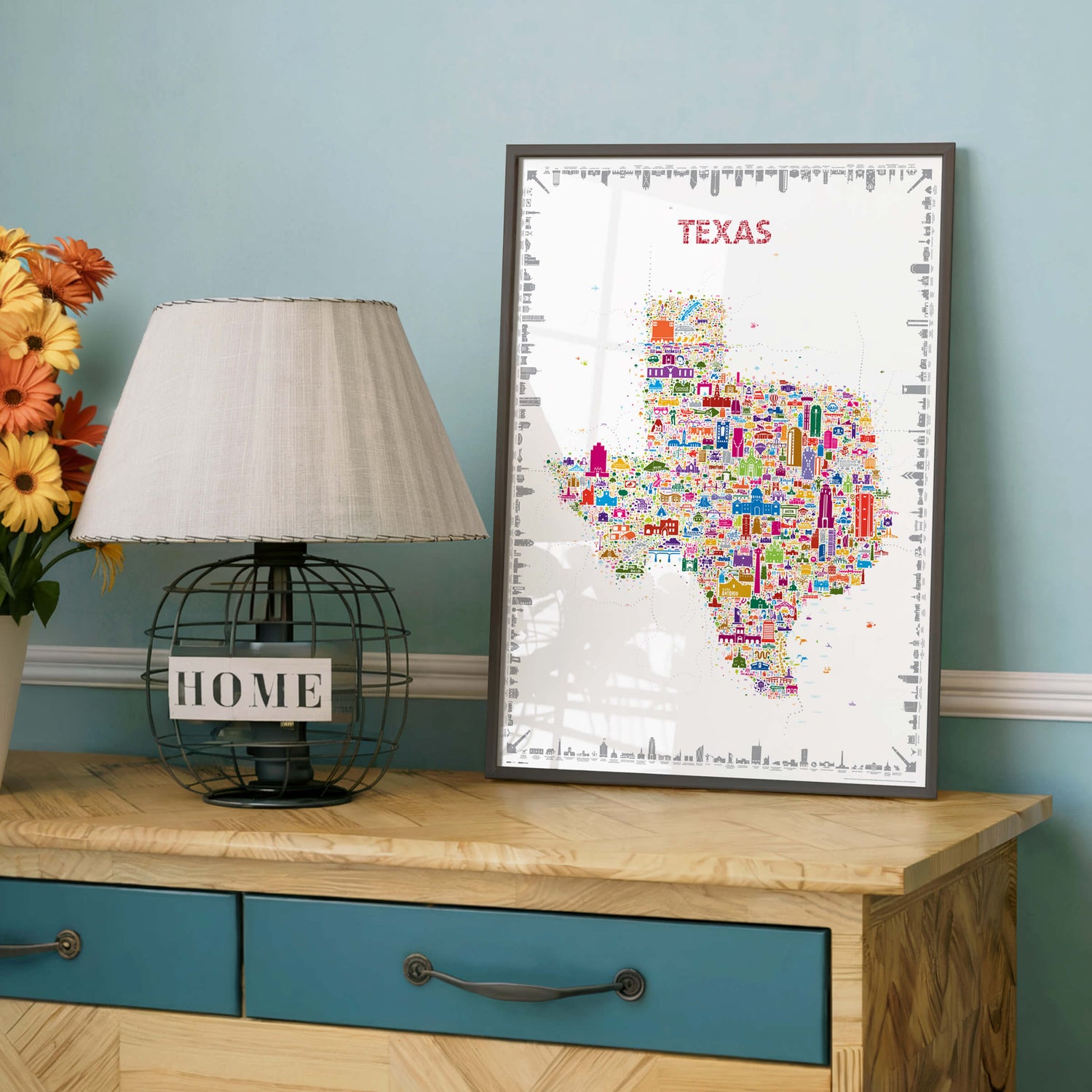 Colorful Texas illustrated map poster in a dark frame displayed on a wooden dresser next to a table lamp and flowers.