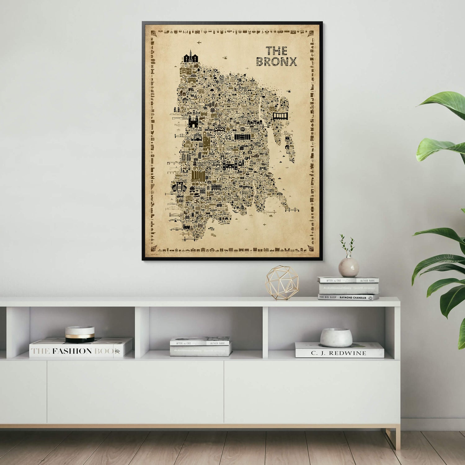 Framed The Bronx Antique illustrated map poster displayed above a modern white console cabinet with books, sculptural decor, a ceramic vase, and green houseplants.
