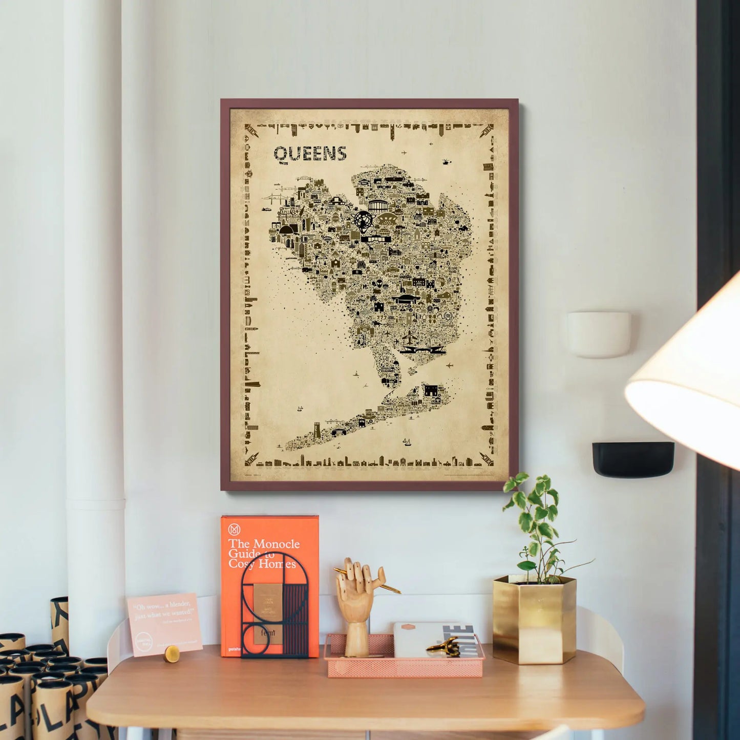 Iconic Queens antique map poster in wooden frame displayed on white wall above modern desk with decorative items and plant