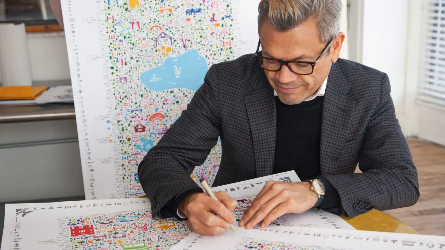Artist hand-drawing colorful illustrated city map poster at desk surrounded by detailed map designs in professional studio