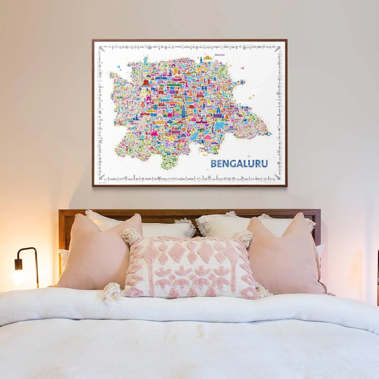 Colorful illustrated Bengaluru city map art print framed above a cozy bed with soft pink and white pillows, creating a warm and modern bedroom aesthetic.