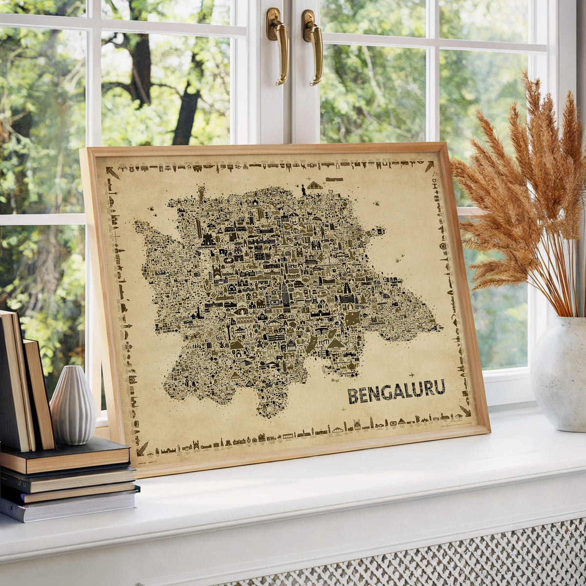 Bengaluru Antique illustrated map poster in a natural wood frame displayed by a bright window with books and dried grasses.