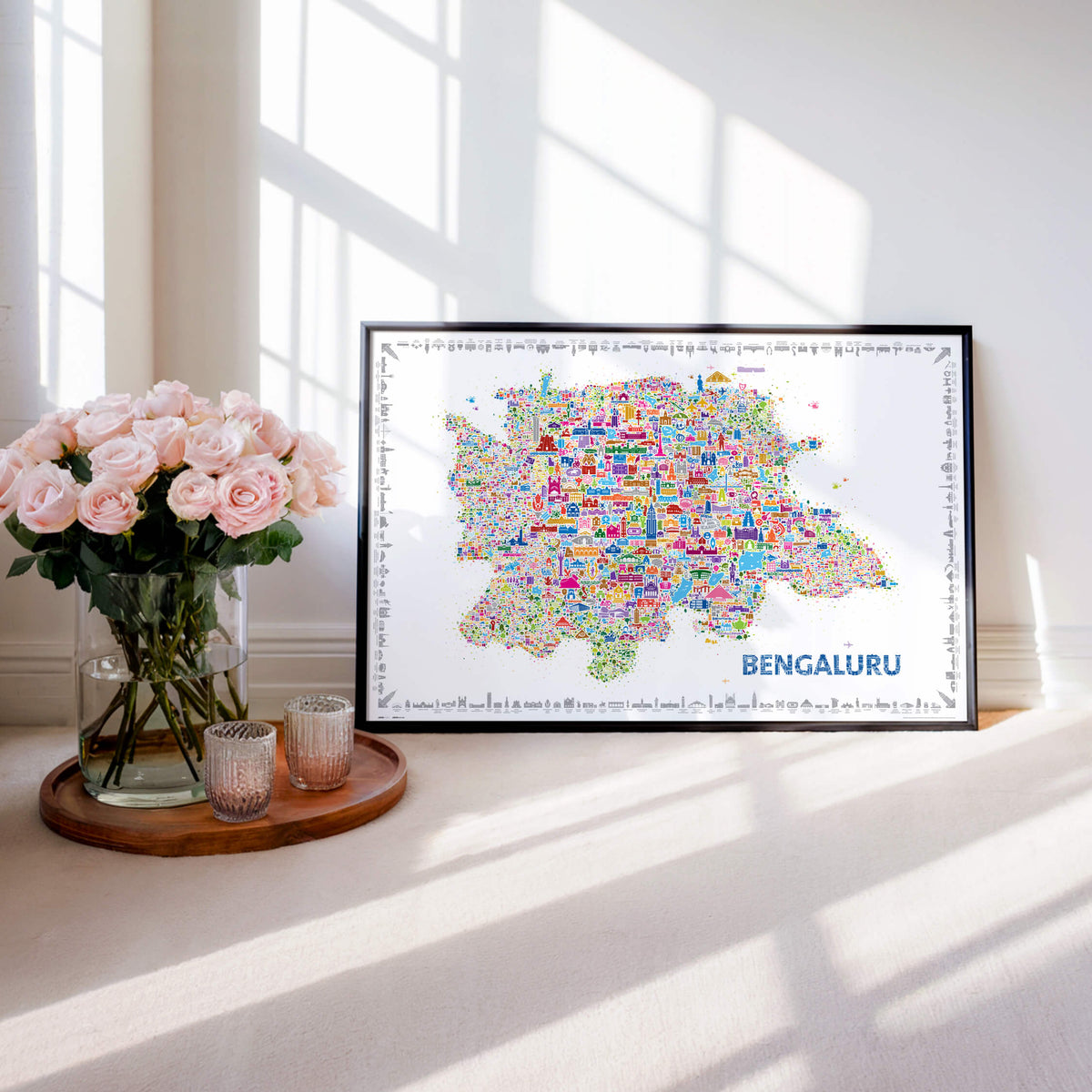Colorful Bengaluru illustrated map poster in a black frame beside pink roses in a bright sunlit interior.