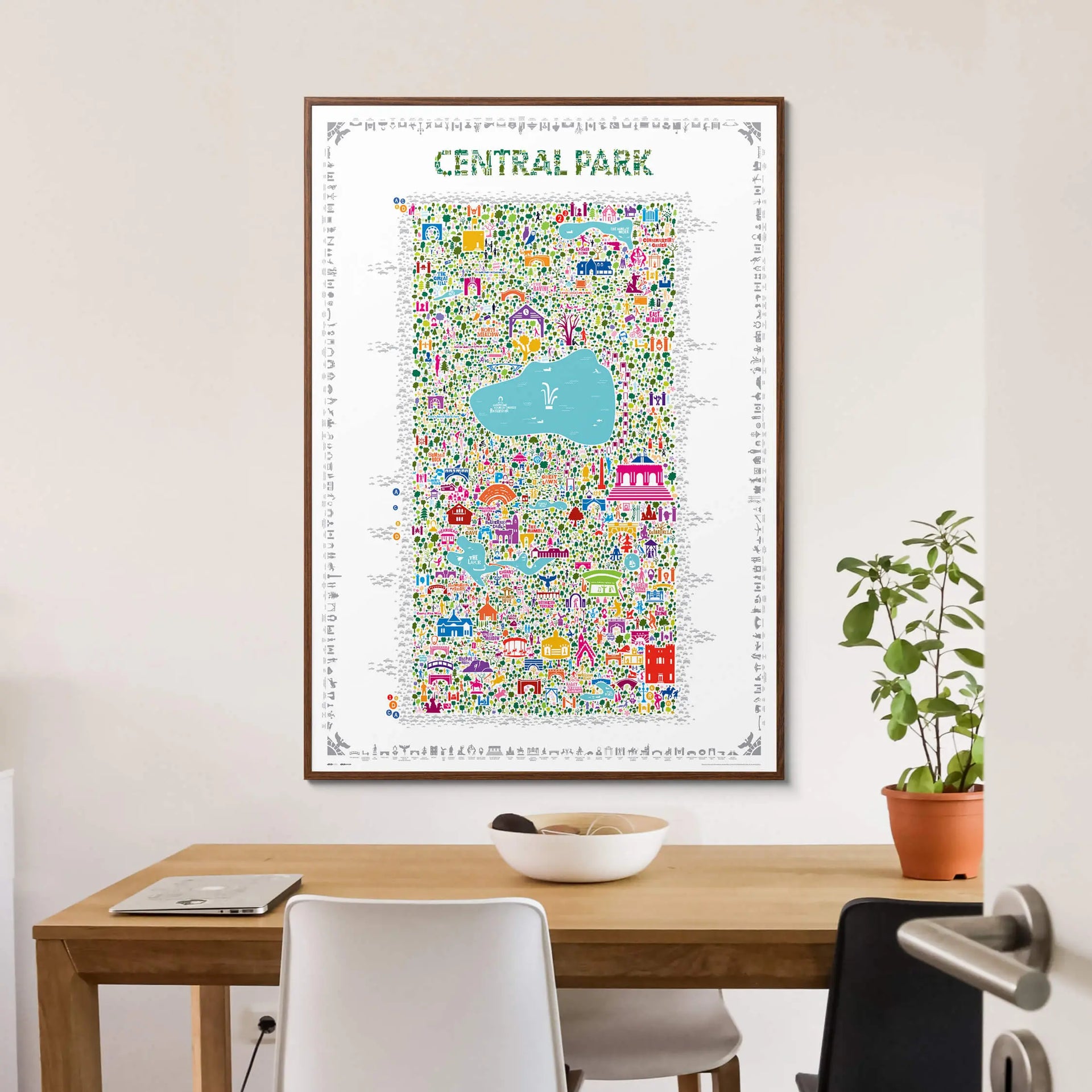 Framed Central Park illustrated map art print hanging above a minimalist wooden dining table with white chairs and indoor plant — modern interior wall art by Alfalfa New York.
