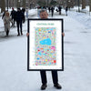 Man standing on a snowy Central Park pathway holding a large framed Central Park illustrated map art print — colorful city landmark poster by Alfalfa New York.