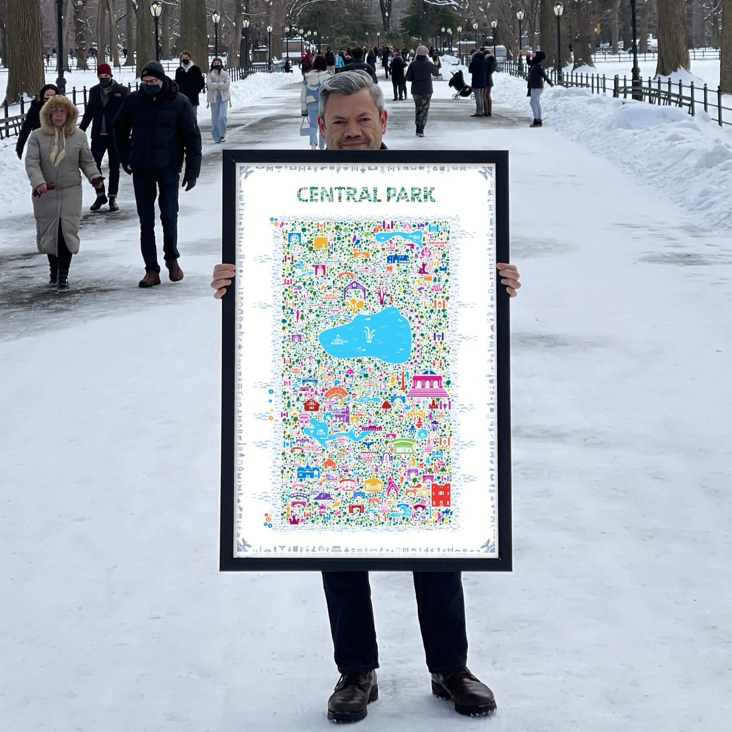 Man standing on a snowy Central Park pathway holding a large framed Central Park illustrated map art print — colorful city landmark poster by Alfalfa New York.