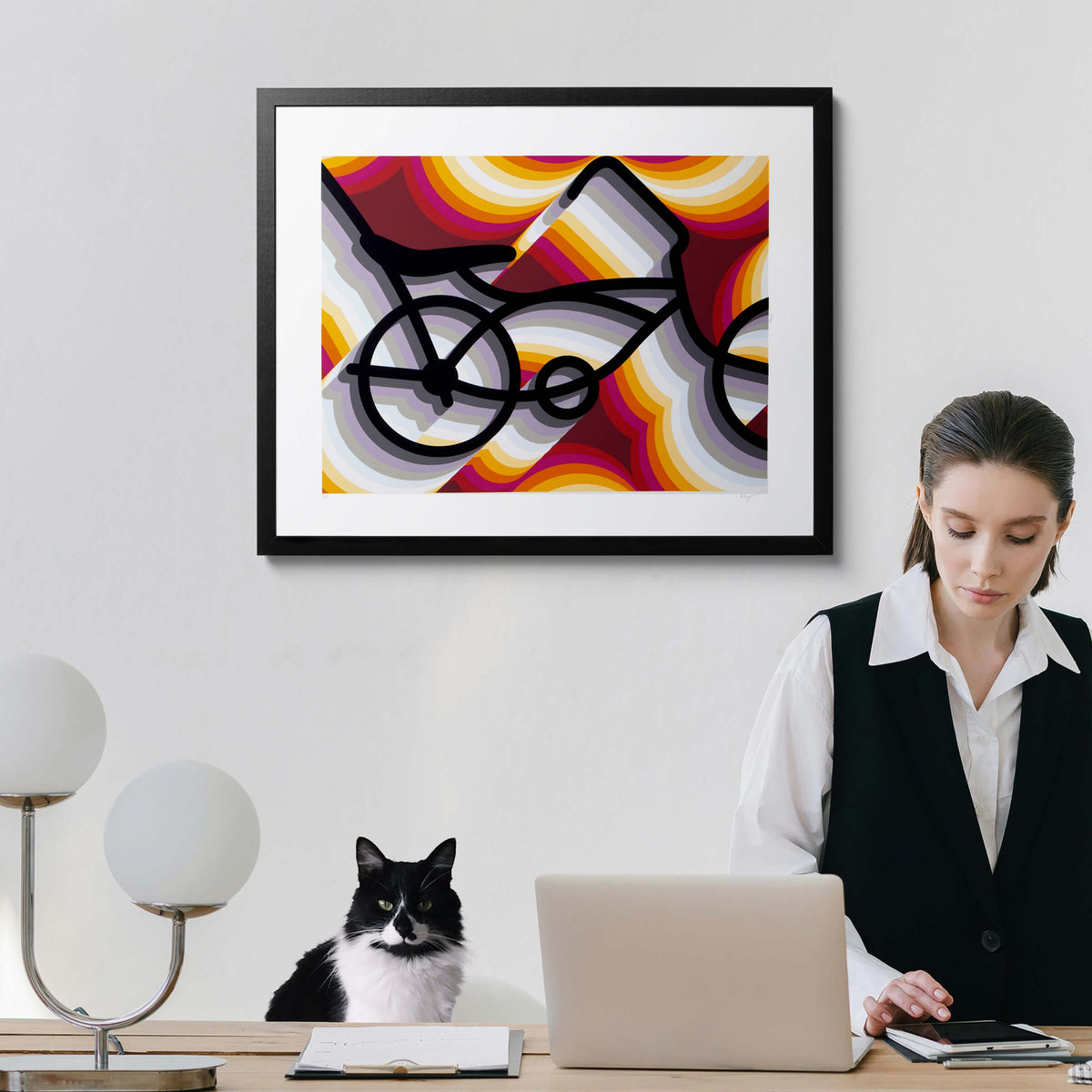 East LA Lowrider Bike screenprint in a black frame on a home office wall, above a desk with a laptop, a woman working, and a black and white cat