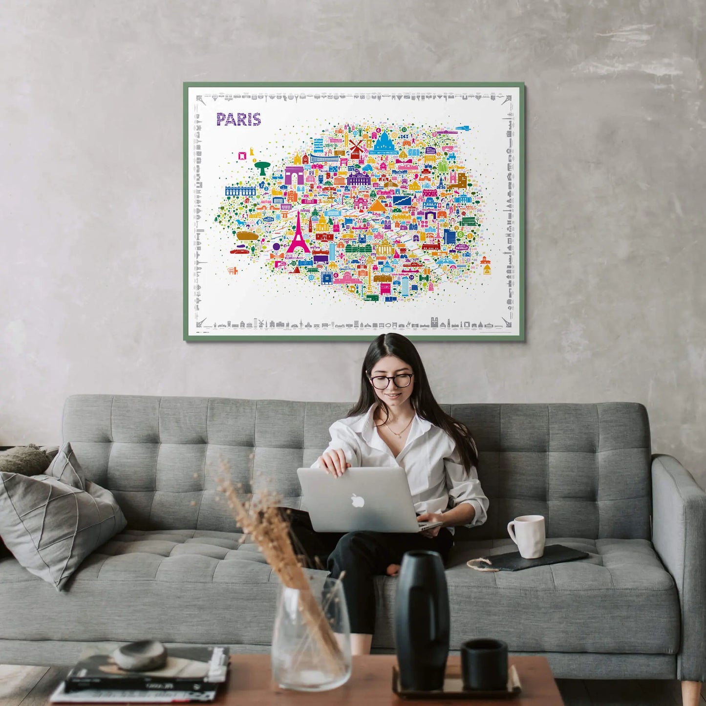 Woman working on laptop on gray sofa beneath colorful Paris illustrated map poster in green frame with modern living room decor