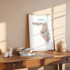Florida map poster in natural wood frame on desk with books and warm sunlight, travel wall art and contemporary home décor