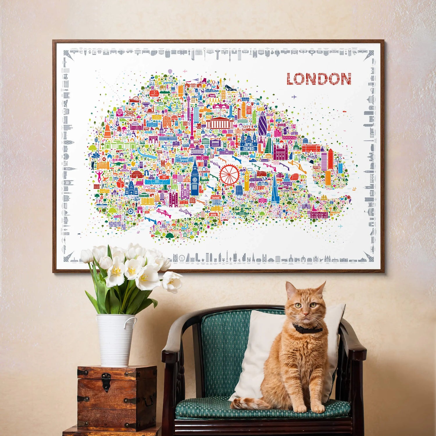 Framed map of London with colorful icons, a cat sitting on a chair below, and a vase with flowers to the left.
