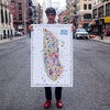 Man standing on a New York City street holding a colorful illustrated Manhattan map poster by Alfalfa New York, showcasing iconic landmarks and modern city art style.
