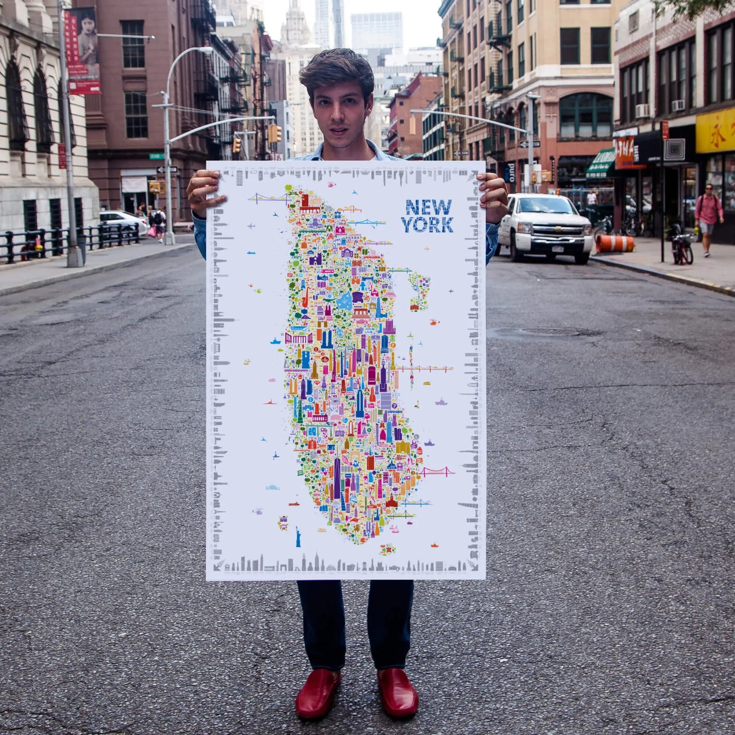 Man standing on a New York City street holding a colorful illustrated Manhattan map poster by Alfalfa New York, showcasing iconic landmarks and modern city art style.

