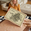 Woman holding a large framed Kolkata Antique illustrated map poster in a natural wood frame while unwrapping the artwork.