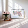 Modern Massachusetts illustrated map poster framed as colorful wall art beside roses and sunlight. Contemporary travel map artwork highlighting Boston, Cape Cod, and New England landmarks in vibrant designer style.