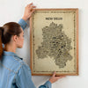 Person hanging a framed New Delhi Antique illustrated map poster in a natural wood frame on a light wall.