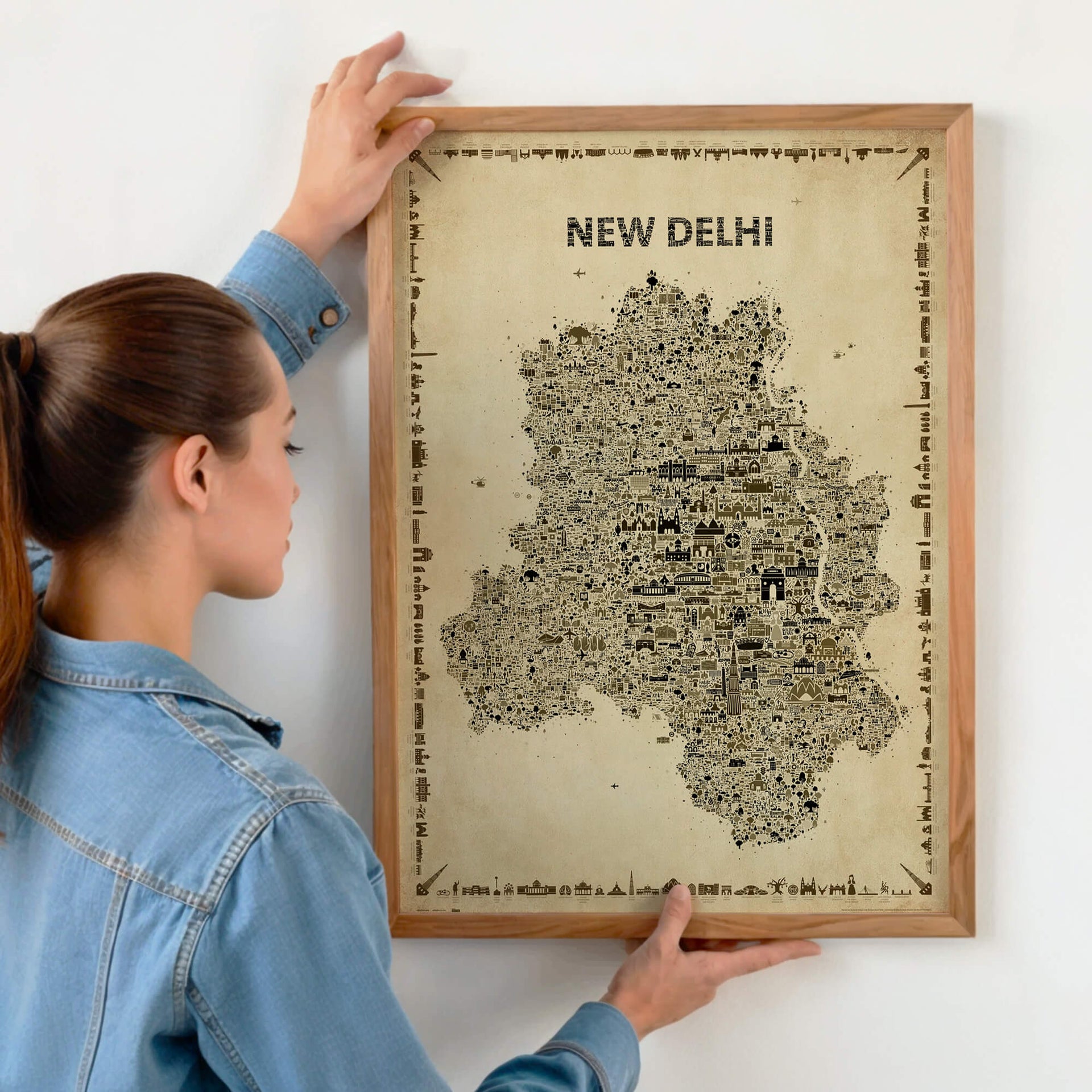 Person hanging a framed New Delhi Antique illustrated map poster in a natural wood frame on a light wall.