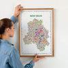 Person hanging a framed colorful New Delhi illustrated map poster in a natural wood frame on a light wall.