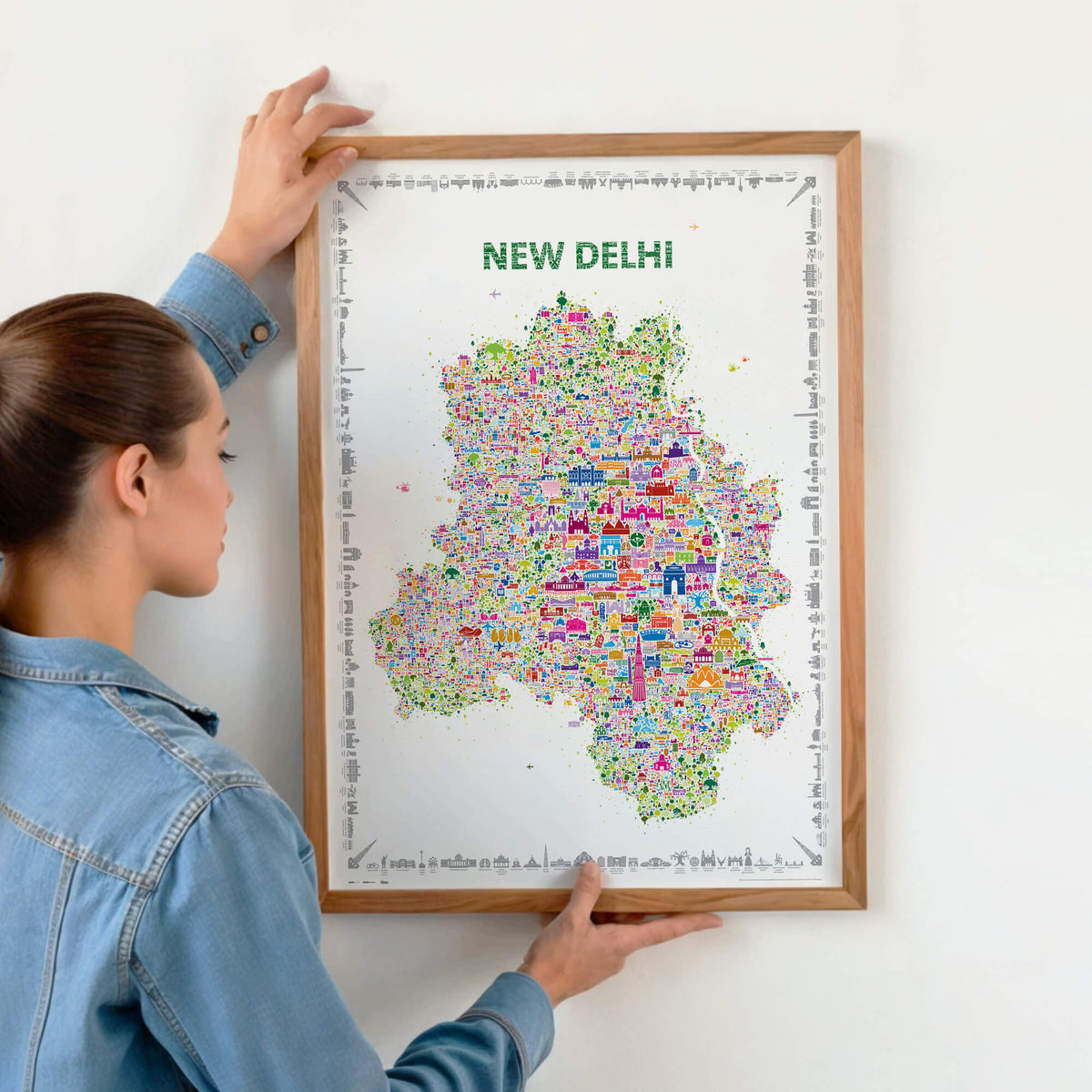 Person hanging a framed colorful New Delhi illustrated map poster in a natural wood frame on a light wall.