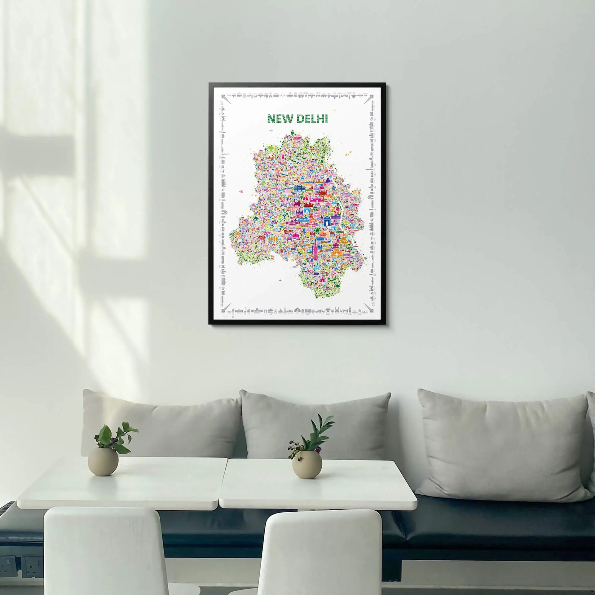 Framed map of New Delhi on a wall above a white sofa with a small table and plants.
