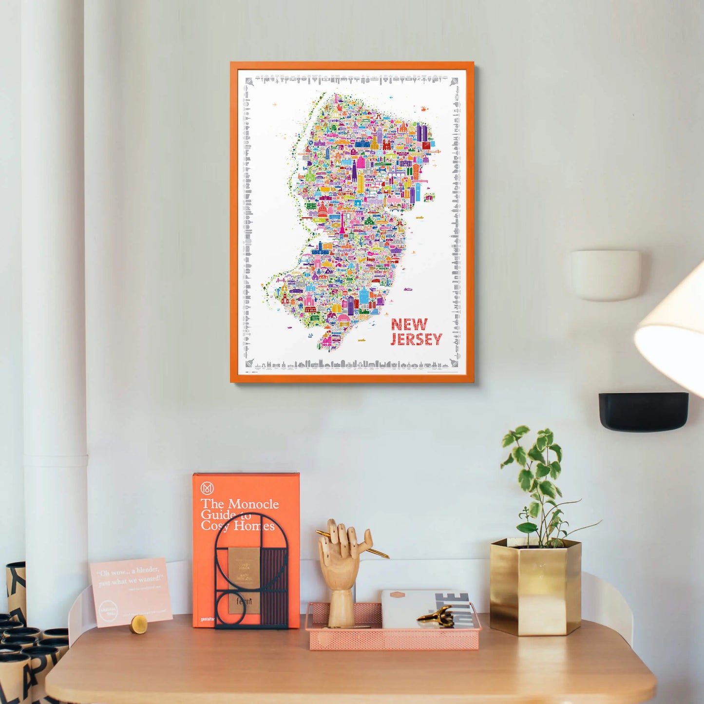 Vibrant New Jersey city map art print with orange frame displayed above a modern study desk — colorful wall decor with books and plants by Alfalfa New York.