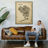 Iconic Queens Poster – Antique Edition in a wooden frame above a brown leather sofa. NYC borough map art in sepia tones styled in a warm, vintage-inspired living room.