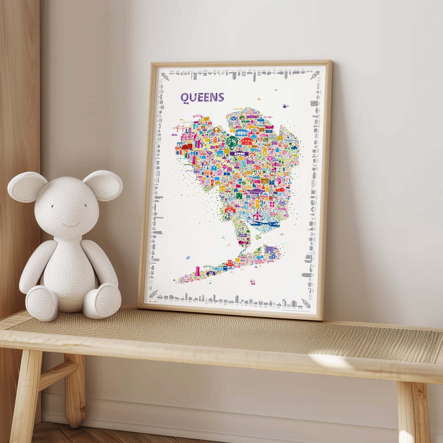 Colorful Queens New York illustrated map poster in a light natural wood frame displayed on a woven bench beside a white teddy bear in a soft neutral kids room or nursery.