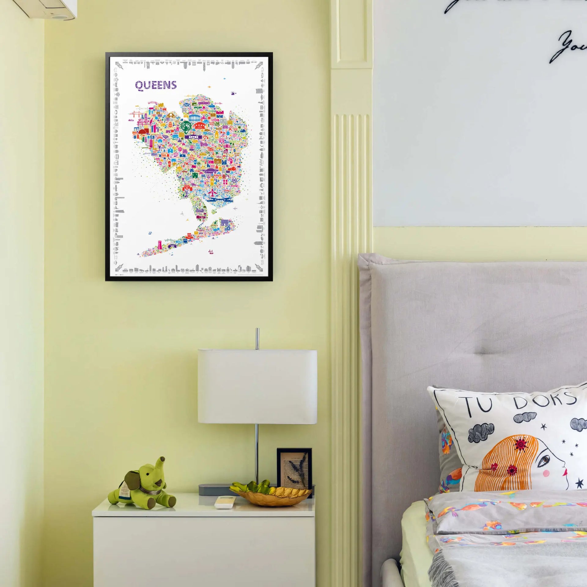 Framed Alfalfa New York Queens illustrated map poster hanging above a nightstand with a lamp and plush toy beside a cozy bed in a softly lit bedroom.