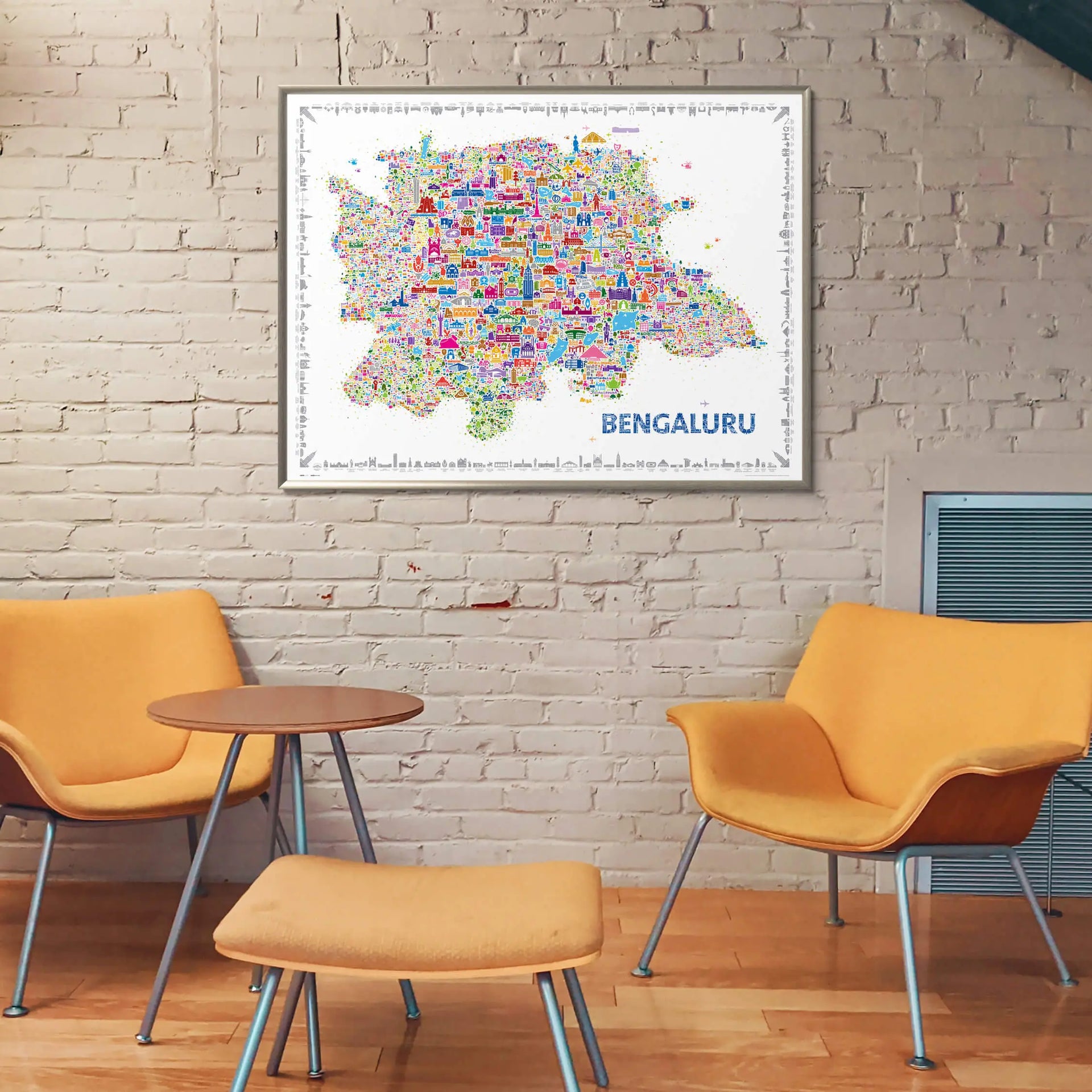 Colorful illustrated Bengaluru city map framed on a white brick wall above modern orange chairs and a wooden coffee table in a cozy contemporary room setting.