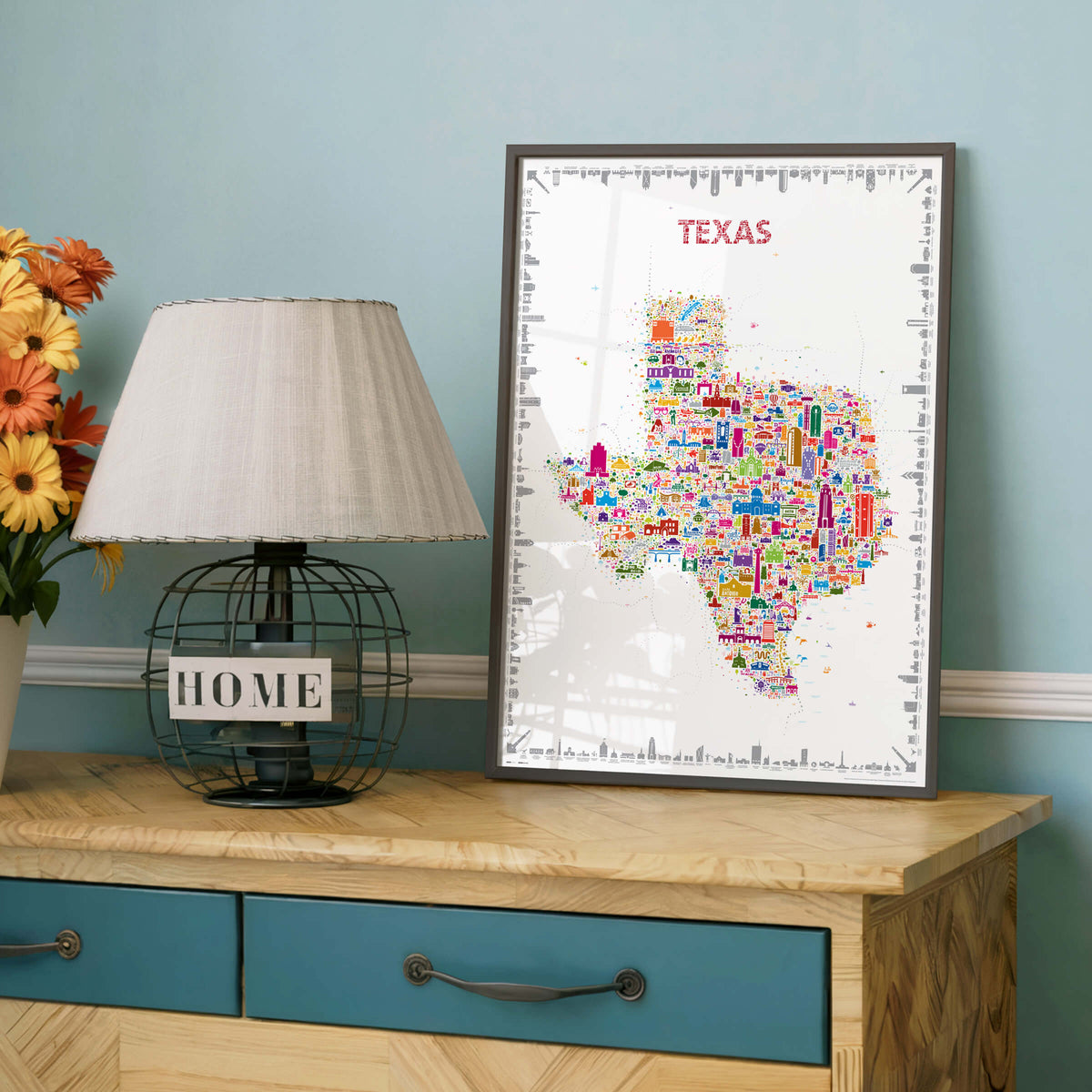 Colorful Texas illustrated map poster in a dark frame displayed on a wooden dresser next to a table lamp and flowers.