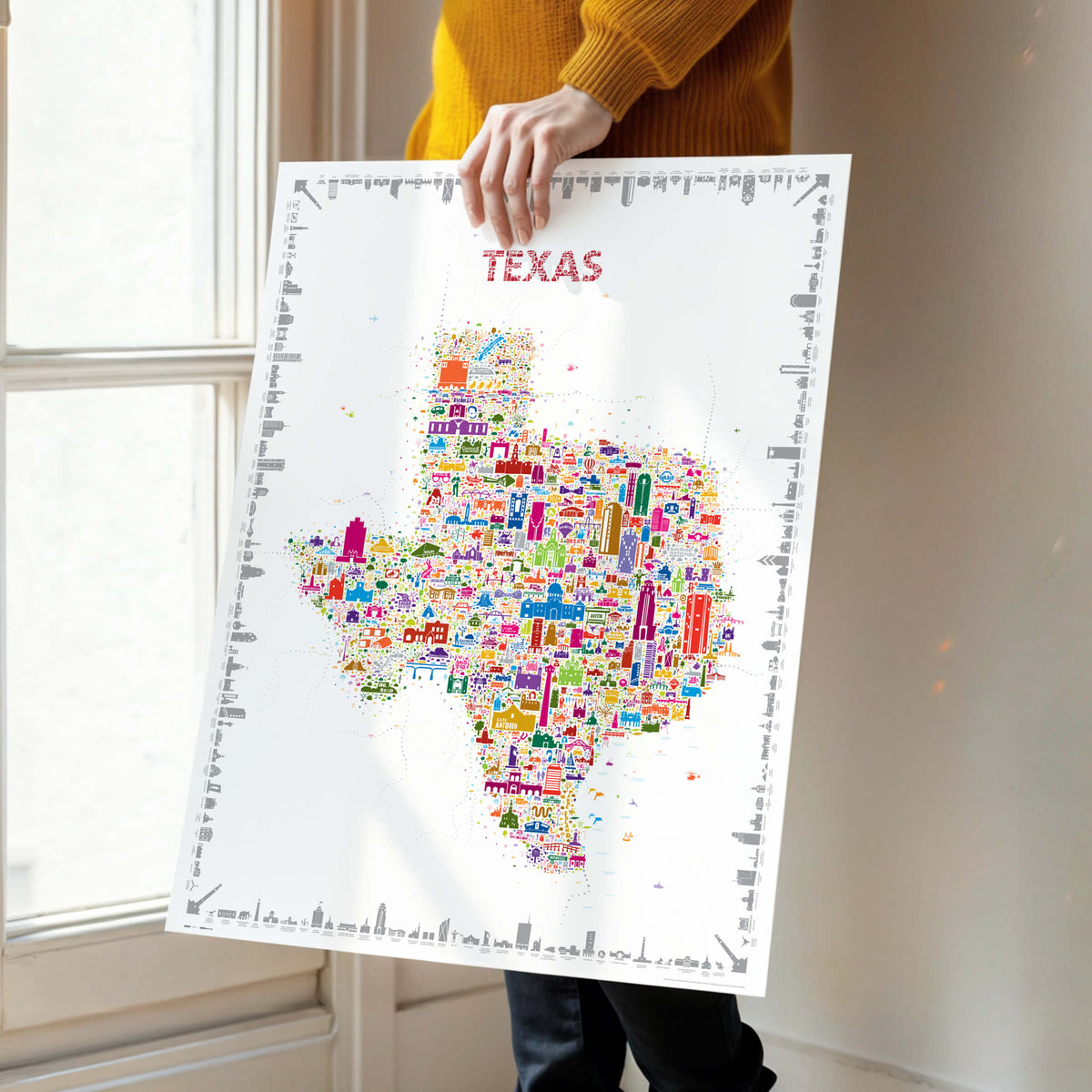 Person holding an unframed colorful Texas illustrated map poster near a window.
