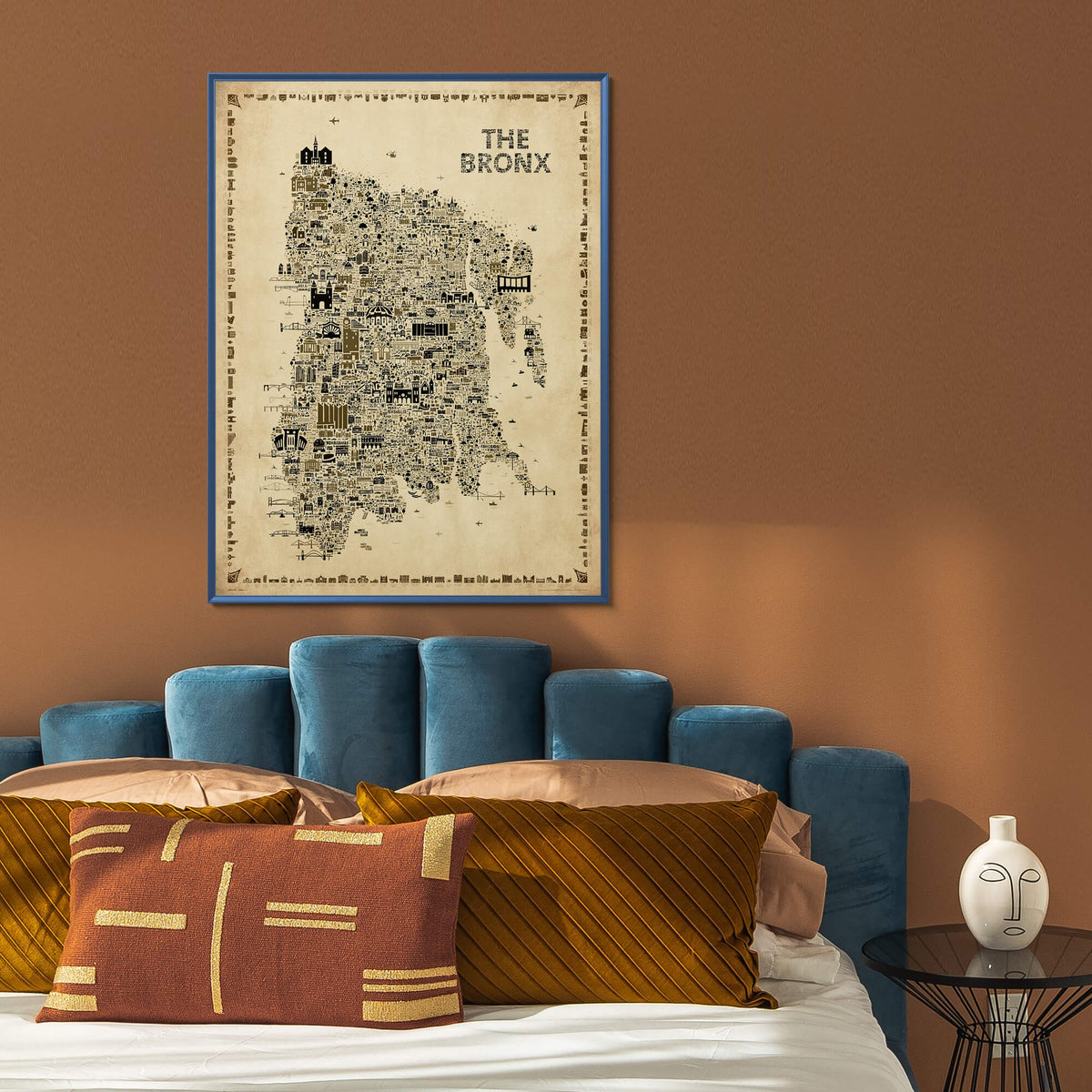 The Bronx Antique illustrated NYC map poster framed above a bed with a blue upholstered headboard, rust and gold pillows, brown walls, and modern bedroom decor.