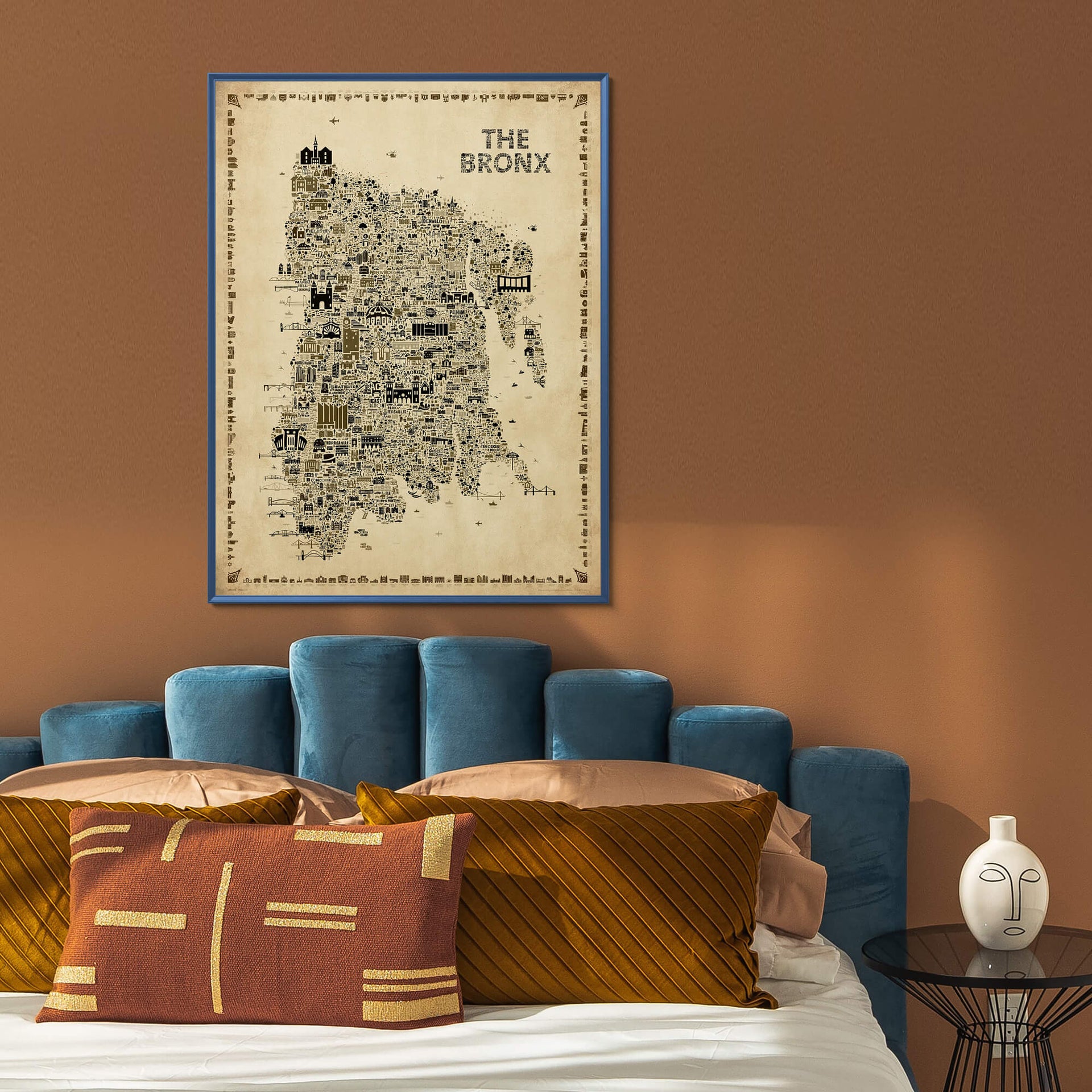 The Bronx Antique illustrated NYC map poster framed above a bed with a blue upholstered headboard, rust and gold pillows, brown walls, and modern bedroom decor.