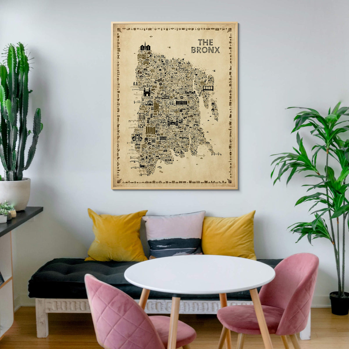 The Bronx Antique illustrated map poster in a natural wood frame hanging above a cozy dining nook with pink chairs, mustard yellow pillows, a black bench, round white table, cactus, and green plants.
