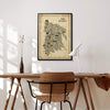 Framed The Bronx Antique illustrated map poster above a minimalist wood desk and chair in a bright home office, with a white task lamp, brown glass vase, dried stems, and sunlight casting shadows across the wall.