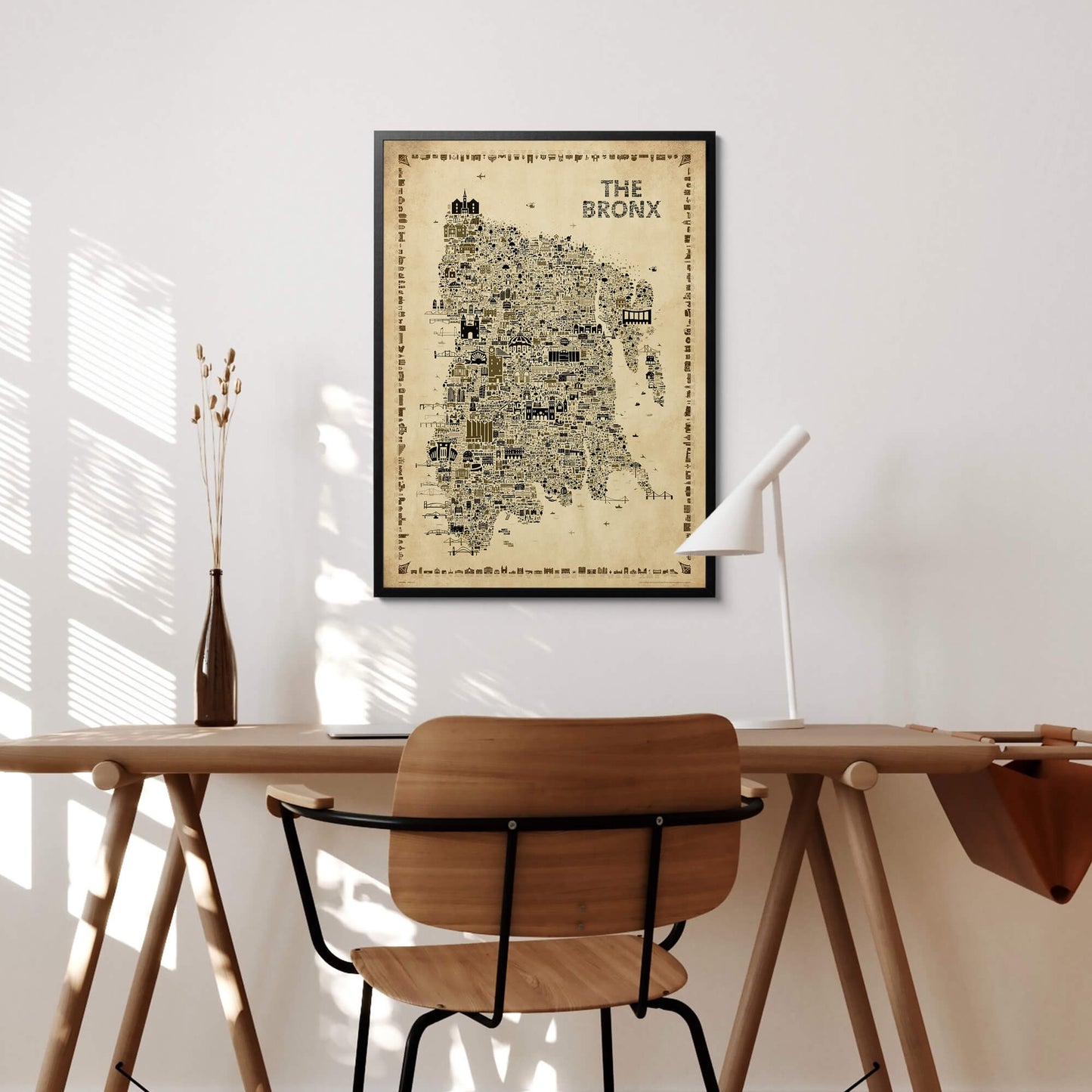 Framed The Bronx Antique illustrated map poster above a minimalist wood desk and chair in a bright home office, with a white task lamp, brown glass vase, dried stems, and sunlight casting shadows across the wall.