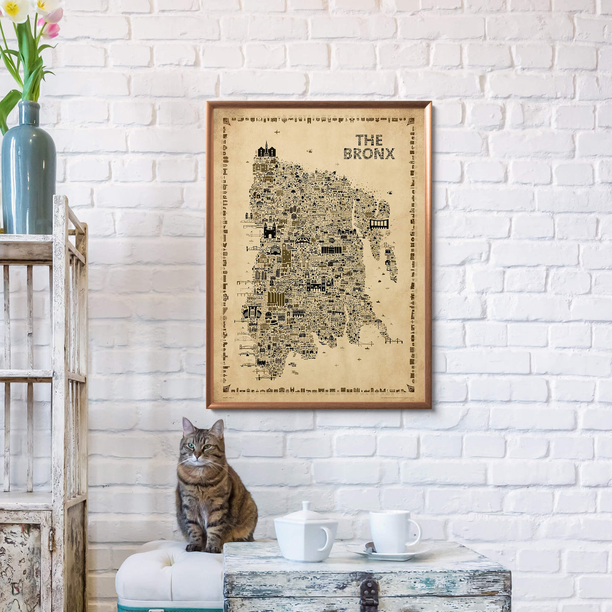 The Bronx Antique illustrated map poster in a warm wood frame hanging on a white brick wall above a small table, with a tabby cat, vintage furniture, flowers, and neutral home decor.